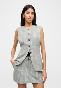 Γυναίκα που φοράει γκρι καρό γιλέκο χωρίς μανίκια με κουμπιά και ταιριαστό ψηλόμεσο καρό φούστα, πόζαροντας με το ένα χέρι στην τσέπη.