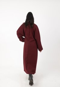 Lang, burgunderfarvet uldfrakke med overdimensionerede ærmer, lige snit, bagsydesøm detalje og båret over sorte chunky støvler. Glat tekstur.