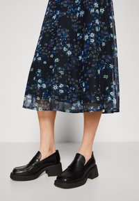 Mocassins noirs à talons épais portés avec une jupe midi en voile bleu marine ornée de petites fleurs blanches et bleues.