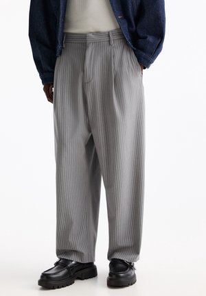 Homme portant un pantalon large à rayures grises, des mocassins noirs épais, une chemise blanche, et une veste en denim foncé, les mains dans les poches.