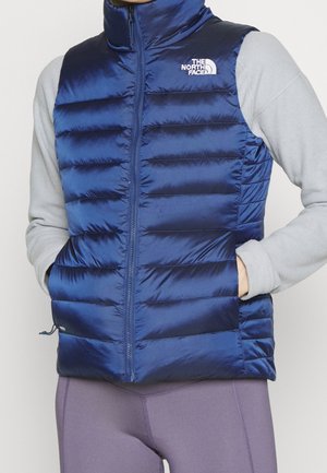 Osoba nosząca niebieską puchową kamizelkę z logo The North Face, szarą bluzkę z długim rękawem i fioletowe legginsy, ręce w kieszeniach kamizelki.
