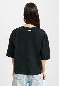 Schwarz oversize T-Shirt mit kurzen Ärmeln, ausgestattet mit einem Rundhalsausschnitt und einem weißen Logo-Detail auf der Rückseite, kombiniert mit hellblauen Jeans.