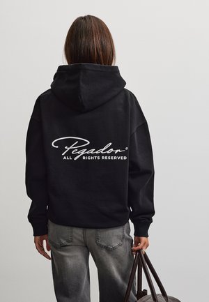 Persona che indossa una felpa nera con cappuccio con testo bianco "Pegador All Rights Reserved" sulla schiena, jeans grigi e tiene manici di una borsa marrone scuro.