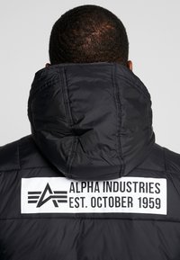 Vista posteriore di una persona che indossa un giubbotto con cappuccio nero con una patch bianca che riporta la scritta "Alpha Industries Est. ottobre 1959" sulla schiena.