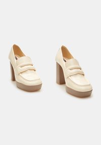 Chaussures en cuir verni crème à bout carré, avec une bride sur le cou-de-pied et un talon épais côtelé. Texture lisse et design minimaliste.