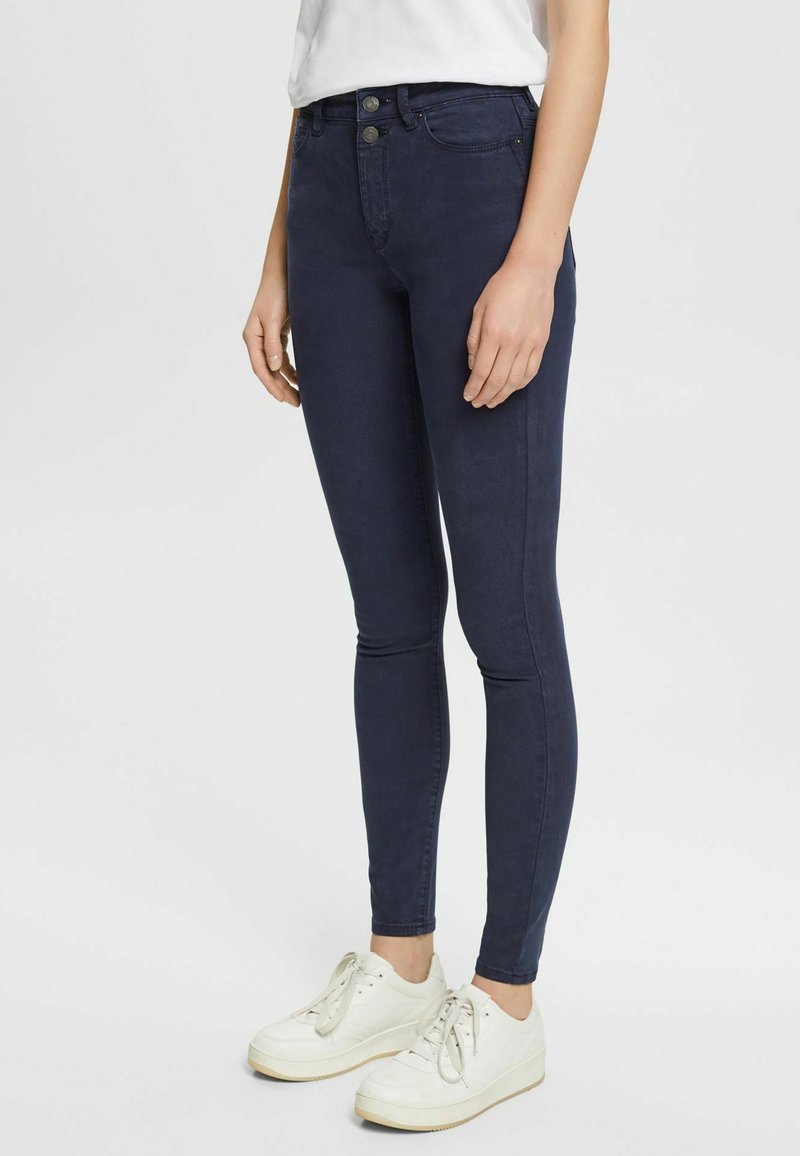Personne portant un jean skinny bleu foncé, des baskets blanches et un haut blanc, debout sur un fond uni.