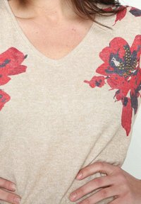 Beżowy sweter z dzianiny z czerwono-nawigacyjnym wzorem kwiatowym, dekoltem w kształcie litery V oraz teksturowanym materiałem z rozrzuconymi ozdobami.