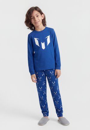 Dreng smilende iført blå langærmet trøje og matchende bukser med hvide grafiske logoer samt grå tøfler på en ensartet hvid baggrund.