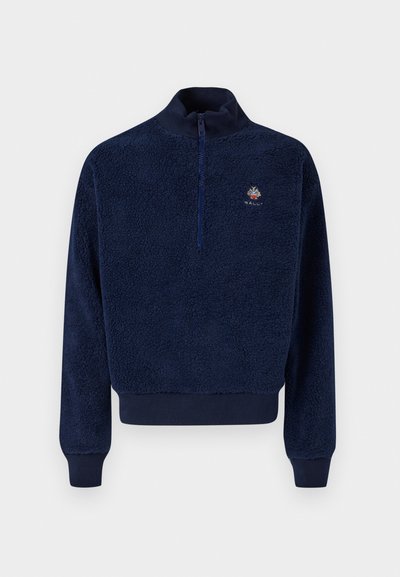 Tums zilas fleece džemperis ar augstu apkakli, priekšējo rāvējslēdzēju un ribotiem aproču galiem, kuram ir mazs izšūts logo uz krūšu apgabala.
