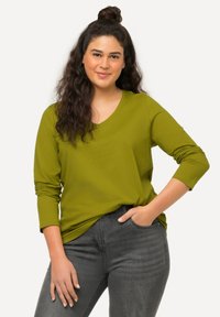 Junge Frau mit langen Locken in einem grünen Langarmshirt und grauen Jeans, die mit einer Hand in der Tasche steht, vor einem hellen Hintergrund.