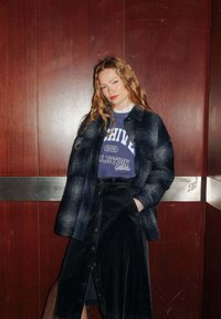 Manteau en denim à carreaux bleu marine avec des accents clairs, porté sur un sweat-shirt graphique bleu, assorti à une jupe en velours côtelé noire avec des boutons.