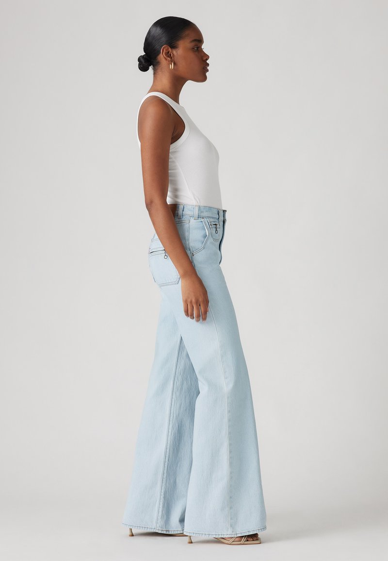 Jeans larges bleu clair en denim, présentant une taille haute et des détails de poches à l'avant. Mannequin portant un top sans manches blanc.
