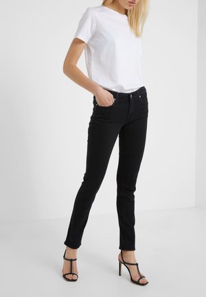 Mujer con una camiseta blanca de manga corta lisa, jeans ajustados negros y sandalias negras de tacón alto, de pie con una mano en el bolsillo.