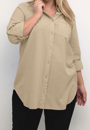 Persoon die een beige overhemd met opgerolde mouwen en een zwarte broek draagt, staand tegen een effen lichte achtergrond.