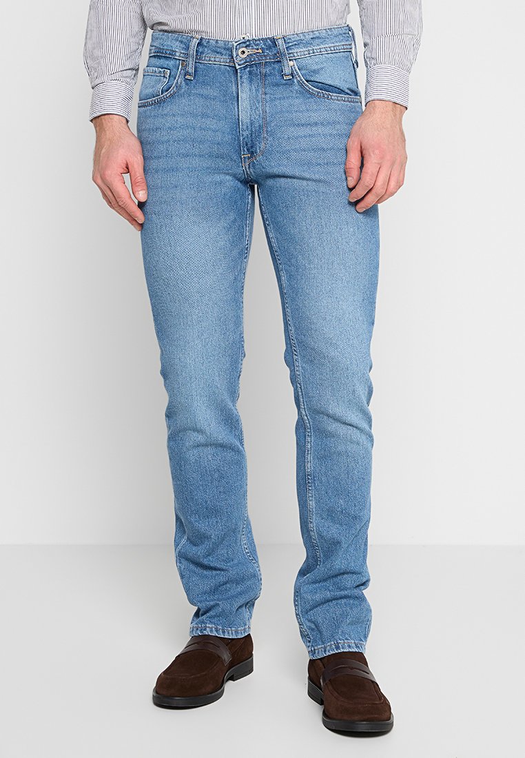 Jeans de mezclilla azul claro con corte slim, que cuentan con cinco bolsillos, un cierre de cremallera y un sutil desvanecimiento. Combinados con zapatos de ante marrón.
