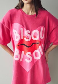 T-shirt in cotone oversize di colore rosa con una grande grafica a forma di cuore, accompagnata dal testo "BISOU" in bianco e un'illustrazione di una labbra rosse al centro.