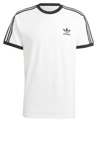 Bílá bavlněná tričko s černým žebrovaným límcem a třemi černými pruhy na rukávech. Obsahuje malé černé logo Adidas na hrudi.