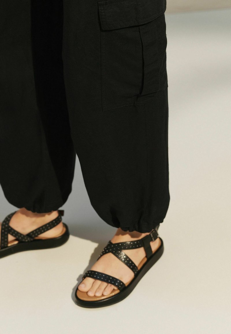 Personne portant un pantalon noir à revers et des sandales noires cloutées sur un sol clair, montrant les jambes et les pieds.