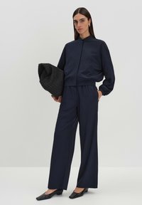 Marineblauwe cropped jasje en wijde broek, beide van een gladde stof. Model houdt een zwarte textured tas vast en draagt slanke zwarte schoenen.