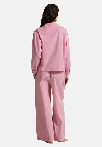 Femme debout, pieds nus, de dos, portant un haut à manches longues rayé verticalement rose et blanc et un pantalon à jambes larges.