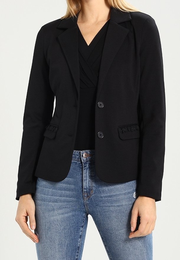 Femme portant un blazer noir ajusté à deux boutons sur un haut noir et un jean bleu clair.
