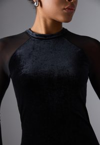 Haut en velours noir avec des manches en résille noires, présentant un col rond et une texture lisse, mettant en valeur une silhouette ajustée.