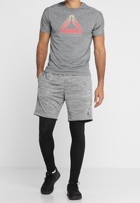 Šedé sportovní tričko s barevným trojúhelníkovým logem, v kombinaci se šedými šortkami a černými stahovacími legínami, doplněné černými sportovními botami.