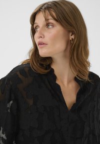 Juoda gėlėta palaidinė, pagaminta iš permatomo audinio, pasižyminti tekstūriniu gėlių raštu, V formos iškirpte ir laisvomis rankovėmis, rodoma kampu.