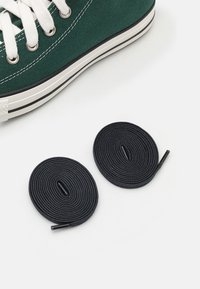 Πράσινο καμβά παπούτσι αθλητικού τύπου με λευκές κορδόνες, διαθέτοντας στρογγυλή μύτη και μαύρη καουτσούκ σόλα. Δύο μαύρες ωοειδείς κορδόνες με άκρες δίπλα στο παπούτσι.