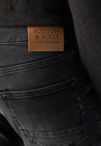 Jeans en denim noir avec une pièce en cuir marron étiquetée "Scotch & Soda Amsterdam". Présente une couture décorative en bleu et gris.