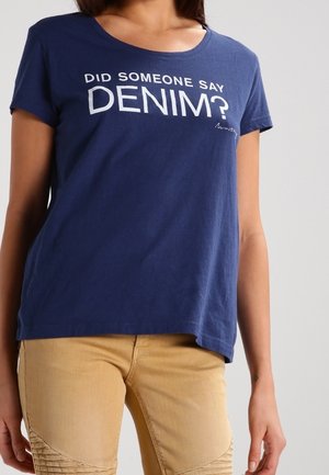 Mujer con camiseta azul marino con el texto "¿Alguien dijo DENIM?" y pantalones beige con parches texturizados en las rodillas, frente a un fondo gris liso.