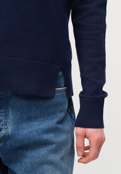 Bras droit et torse d'une personne portant un pull bleu marine avec un ourlet fendu sur le côté et un jean en denim bleu sur un fond neutre.