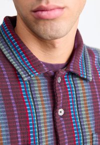 Button-up shirt featuring a plaid pattern in burgundy, green, and blue hues. The collar is lined with contrasting colored stripes.