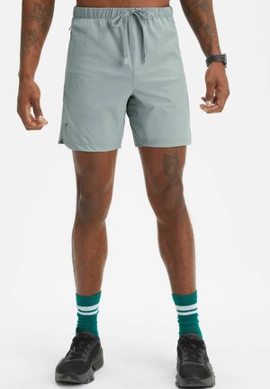 Shorts de bain gris clair avec une taille élastique et un cordon de serrage, poches latérales et coupe décontractée. Portés avec des chaussures de sport noires et des chaussettes turquoise.