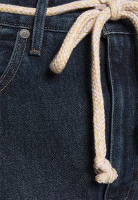 Tummansininen denimkangas, jossa on punottu nauha vaaleanpunaisissa ja keltaisissa sävyissä, solmittuna edessä näkyvillä ompelutiedoilla.