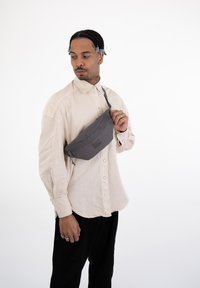 Marsupio grigio realizzato in materiale resistente, con chiusura a zip e tracolla regolabile, indossato sopra una camicia button-up beige chiaro.