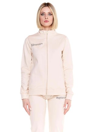 Un mannequin est de face, vêtu d’une veste rose pâle zippée et d’un pantalon assorti, sur fond blanc.