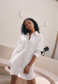 Robe chemise blanche brodée avec des motifs floraux, des manches longues et une fermeture à boutons sur le devant. Le modèle est posé devant un fond aux tons neutres.