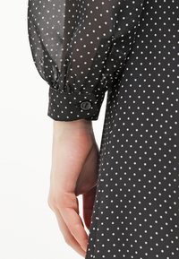 Chemise noire à pois avec des manches transparentes et bouffantes, et un détail de poignet boutonné. Le tissu a une texture lisse avec une apparence légère.