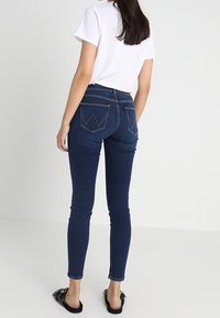 Ändå en tight mörkblå denimbyxor med hög midja. Har två bakfickor med en dekorativ sömnadsmönster. Bärs med en vit t-shirt.