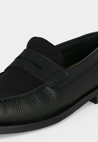 Mocassin noir en cuir texturé avec un design moc toe et un détail de bride sur le dessus, présenté sur un fond clair uni.