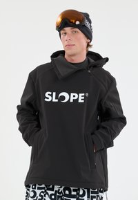 Sort skijakke med grå accenter og høj krave, der har et fremtrædende hvidt "SLOPE" logo. Modellen bærer orange skibriller og mønstrede bukser.