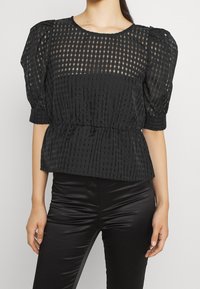 Blusa nera con maniche a palloncino, caratterizzata da un motivo a rete testurizzato, vita aderente e parte superiore in tessuto trasparente con scollatura rotonda.