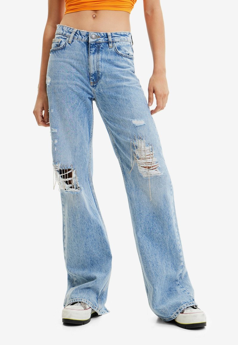 Femme portant un jean large bleu clair déchiré avec des détails en chaîne, des baskets blanches et un top court orange, debout les mains détendues.