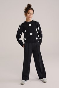 Fekete pulóver nagy fehér pöttyökkel, laza fekete nadrággal párosítva. A ruházat laza szabású, bordázott mandzsettákkal.