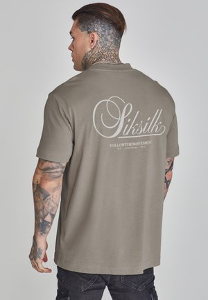 Polo a maniche corte color khaki con una texture liscia. Presenta un grande logo bianco del marchio sul retro e dettagli di tatuaggi visibili sul braccio.