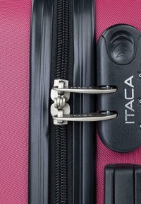 Detalle del zipper de una maleta rosa con dos tiradores de metal plateado, carril de zipper negro y etiqueta de logo. La tela texturizada muestra un diseño duradero.