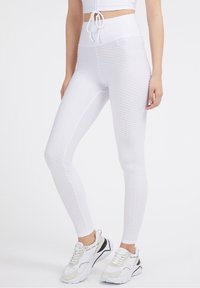 Žena v texturovaných bílých legínách s vysokým pasem, odpovídajícím zkráceném topu se stahovací šňůrkou a robustních bílých teniskách na jednolitém pozadí.