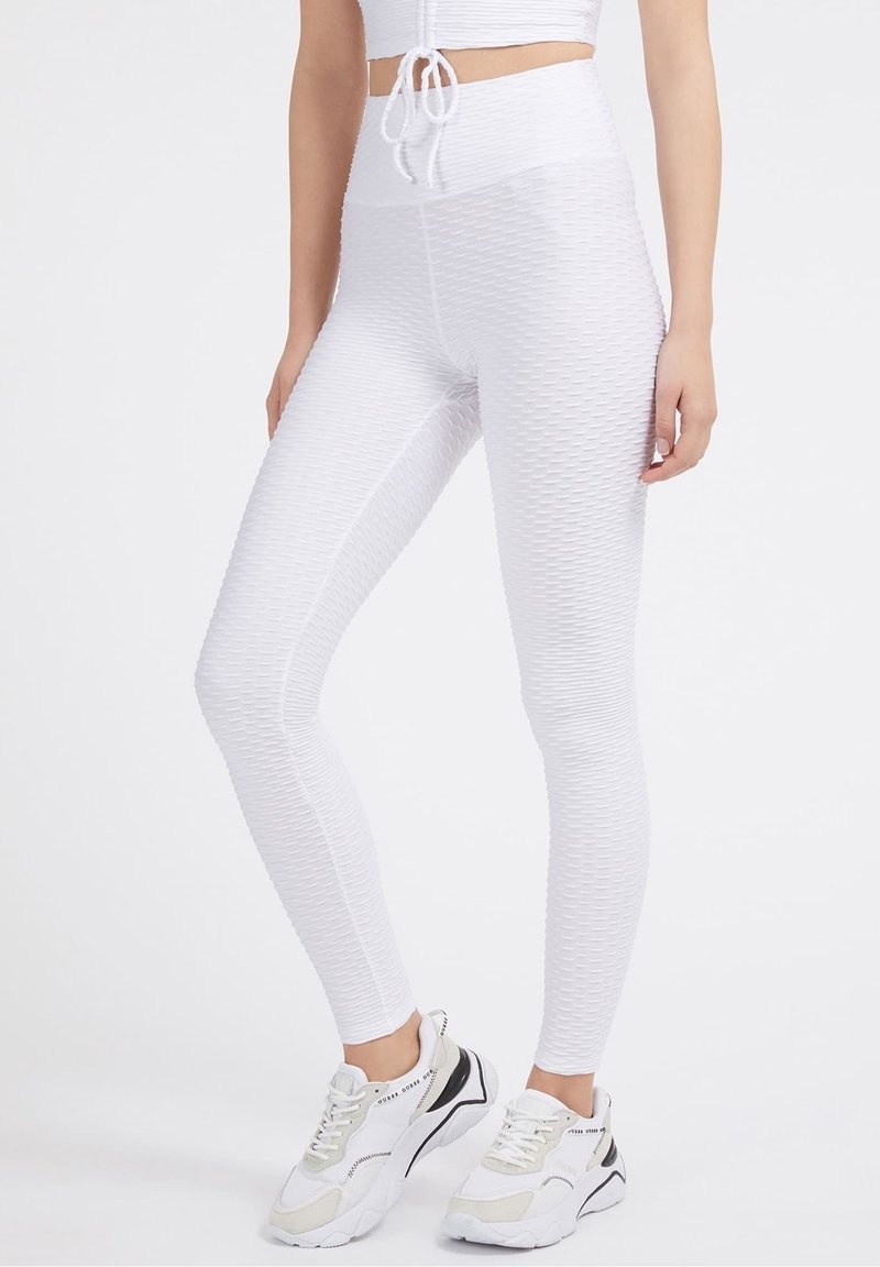 Žena v texturovaných bílých legínách s vysokým pasem, odpovídajícím zkráceném topu se stahovací šňůrkou a robustních bílých teniskách na jednolitém pozadí.
