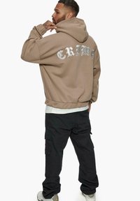 Hoodie oversized marron avec une capuche à cordon, arborant le mot "CRIME" en lettres blanches dans le dos ; associé à un pantalon cargo noir.